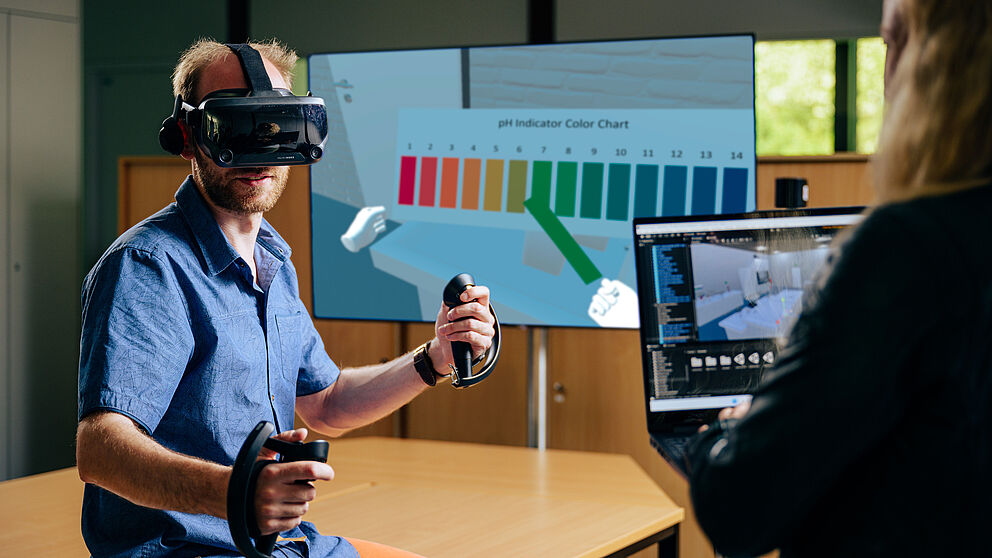 Eine Person nutzt ein VR-Headset. Im Hitergrund sieht man die Sicht der Person im VirtuChemLab.